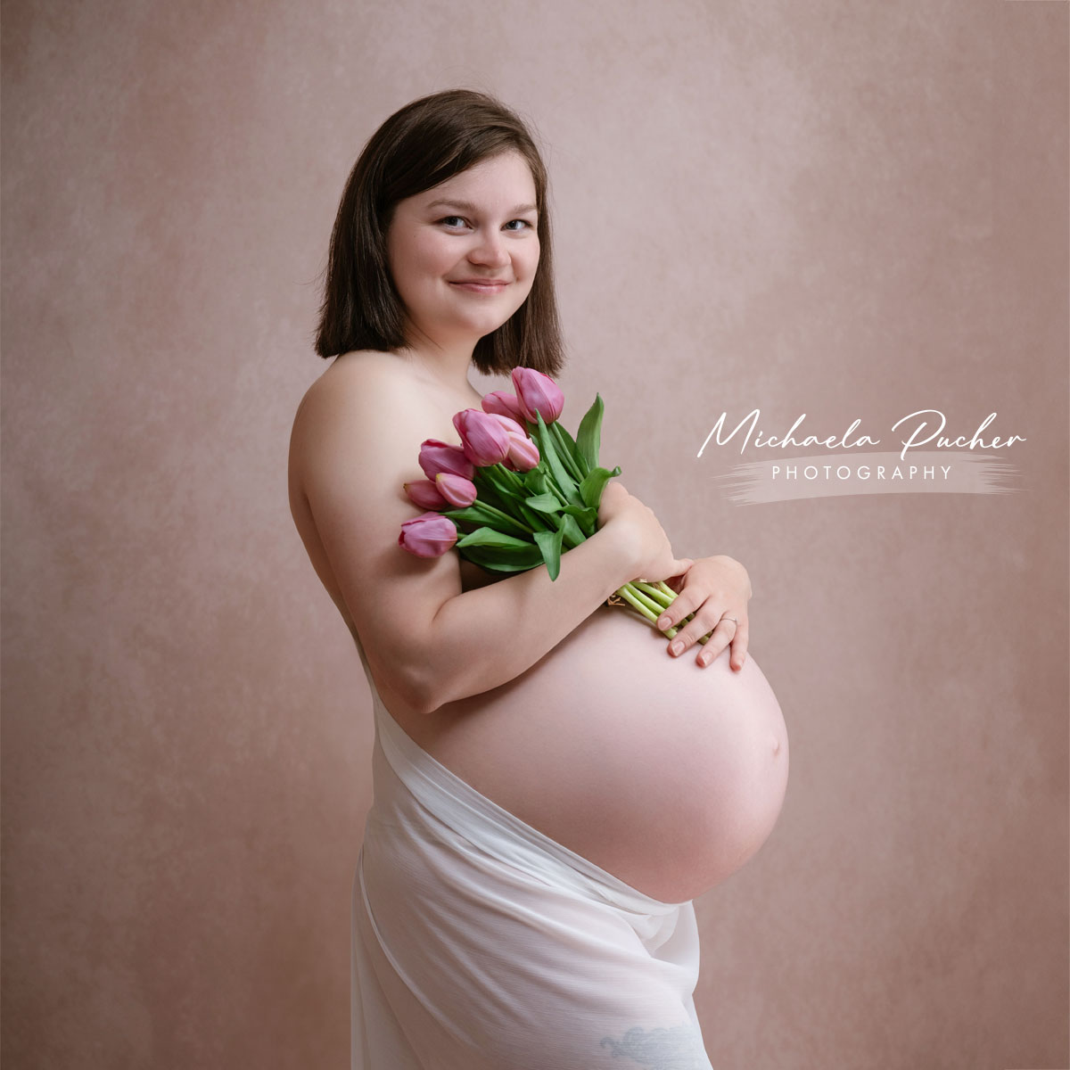 Hochschwangere Frau im Studio, seitlich fotografiert, hält einen Strauß rosafarbener Tulpen vor ihrem Babybauch und lächelt ruhig in die Kamera.